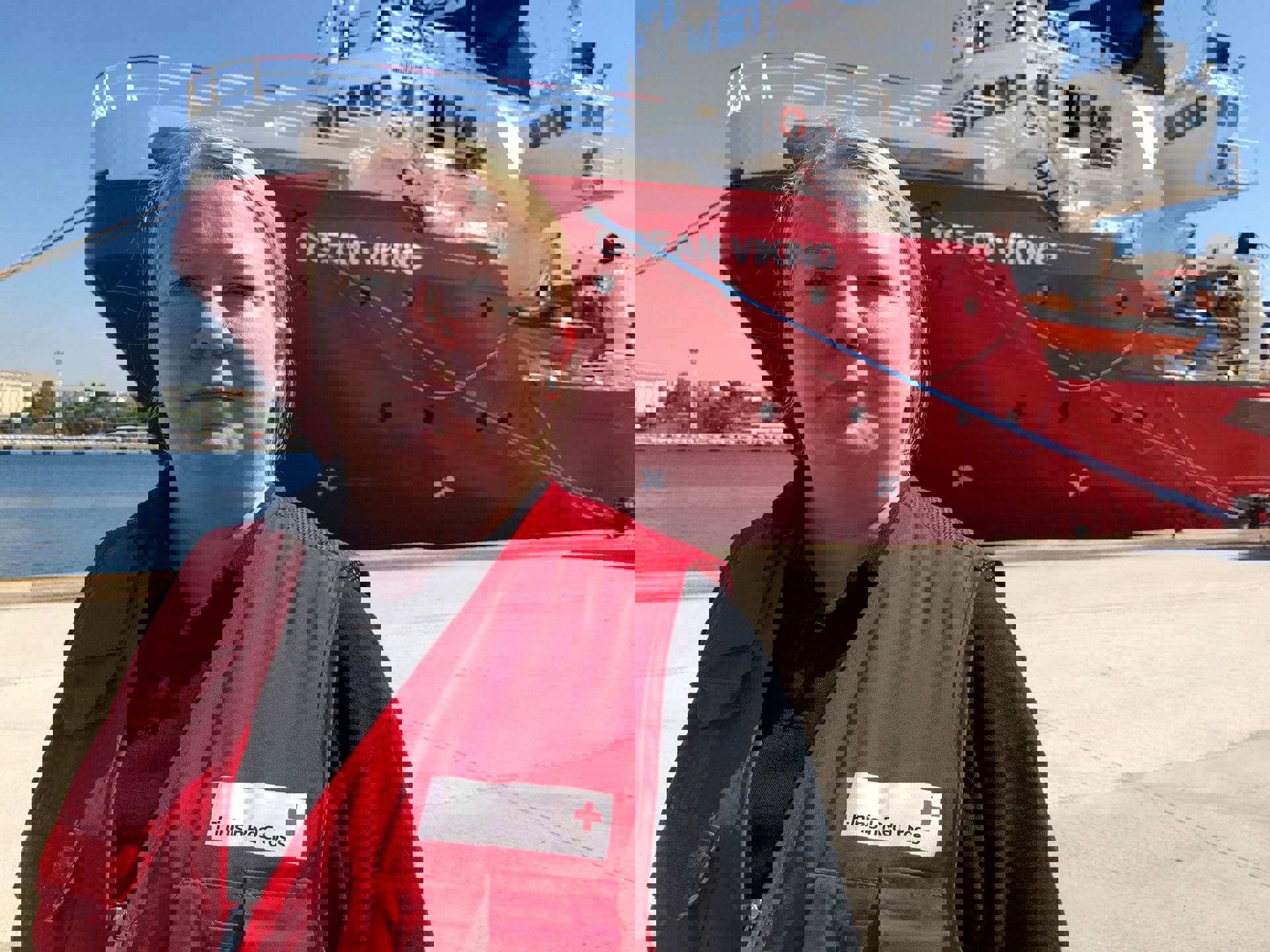 Biståndsarbetare klädd i Röda Korsets arbetsdräkt står framför ett fartyg på hamnområdet.
