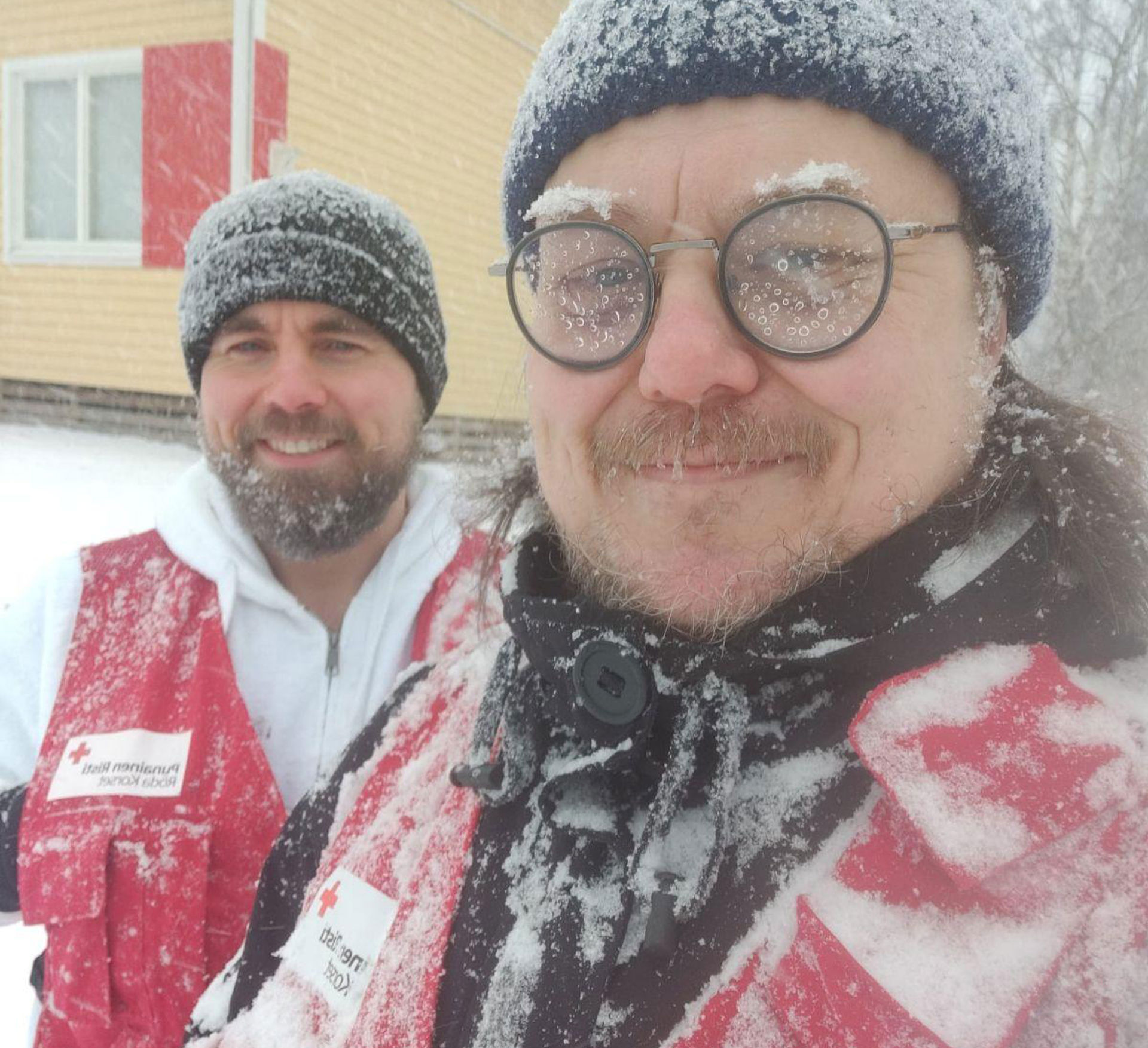 Teemu Hiilinen och Vesa Vahermaa står leende i mycket snöigt väder, iklädda Röda Korsets frivilligvästar.