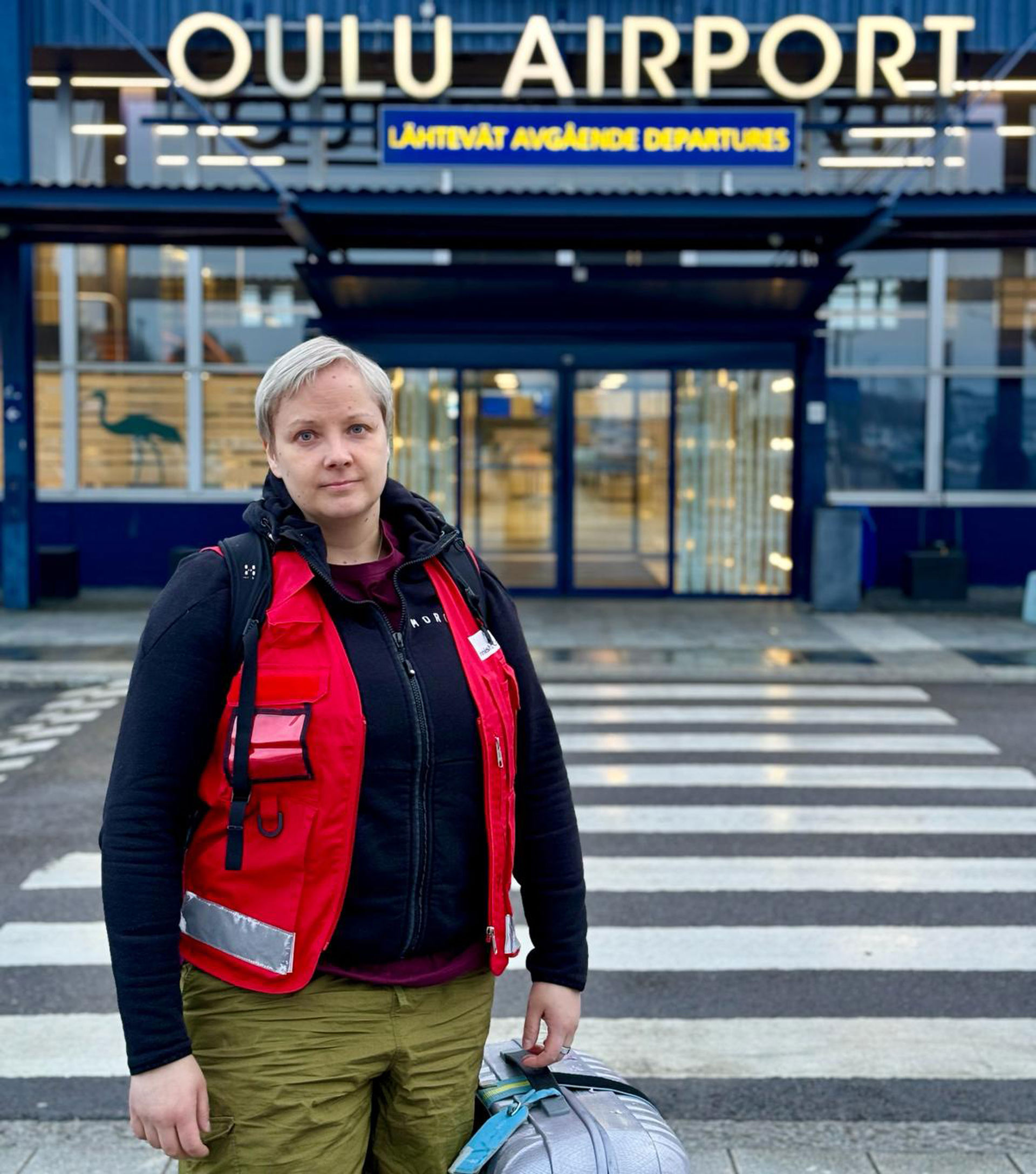  Anni Walli står i Röda Korsets arbetskläder framför Uleåborgs flygplats med en resväska i handen.