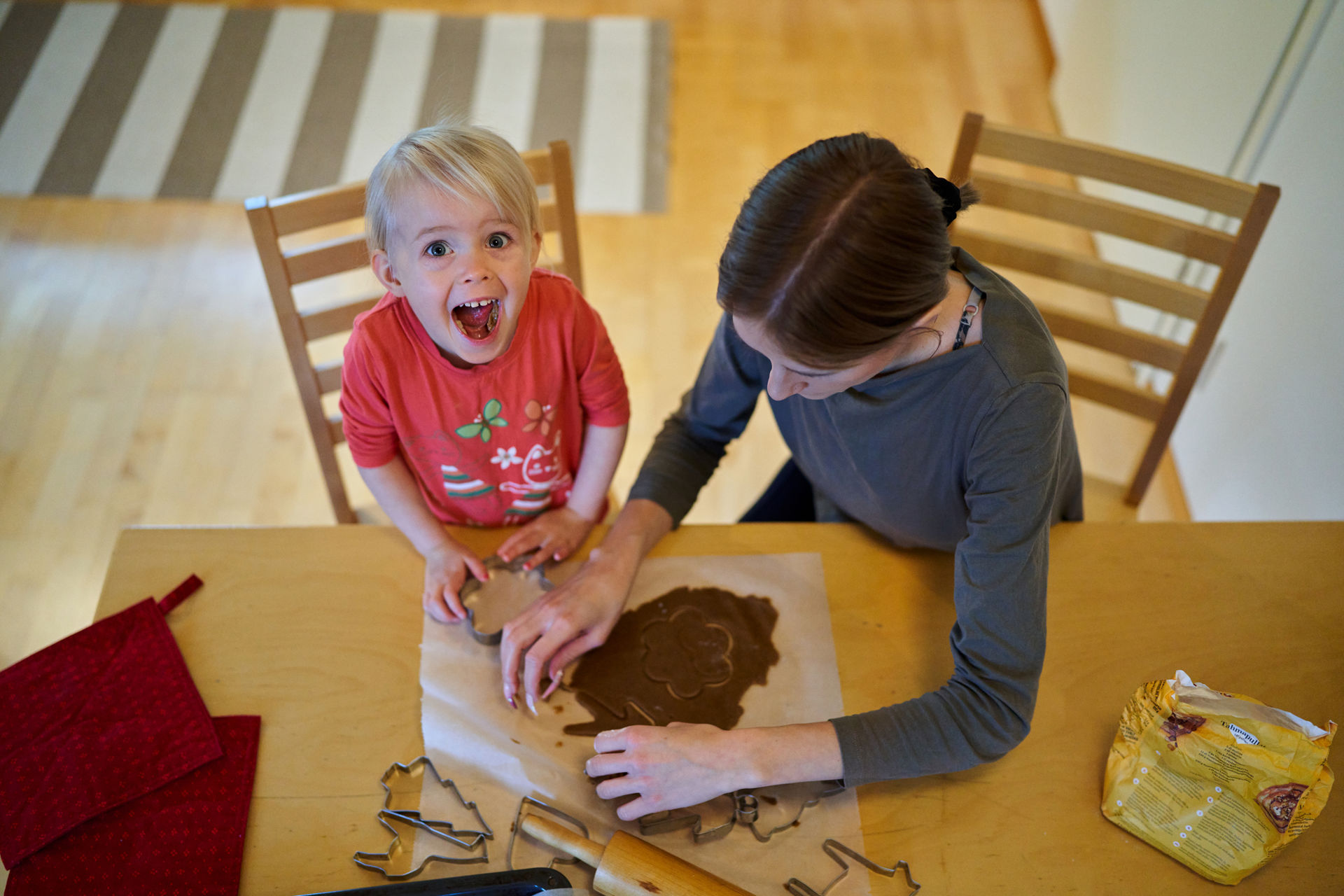 En ung flicka tittar på kameran med ett glatt uttryck. Hon sitter vid bordet med sin mamma och bakar pepparkakor.