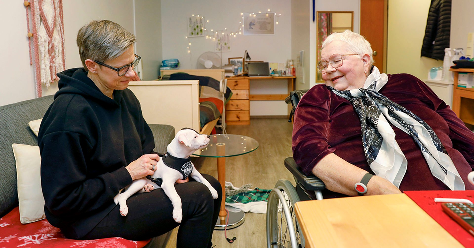 En frivillig och en äldre person sitter vid bordet och ler. Frivilligaren har en liten hund i famnen.