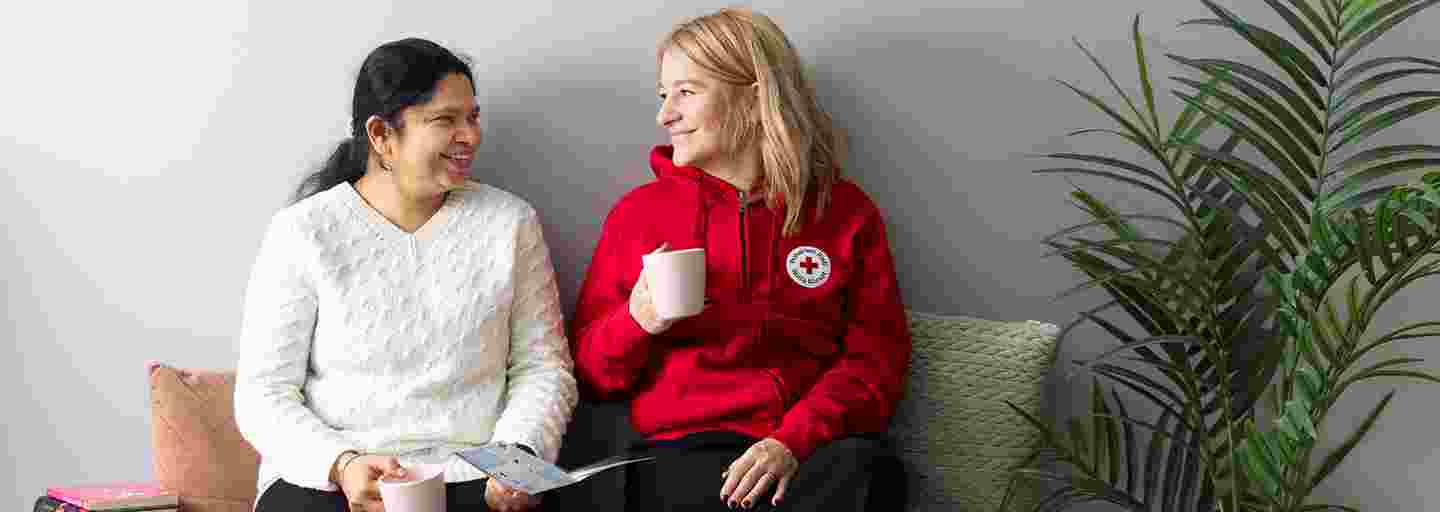 Två leende personer sitter på en soffa med Röda Korsets broschyr och en kaffekopp i handen.